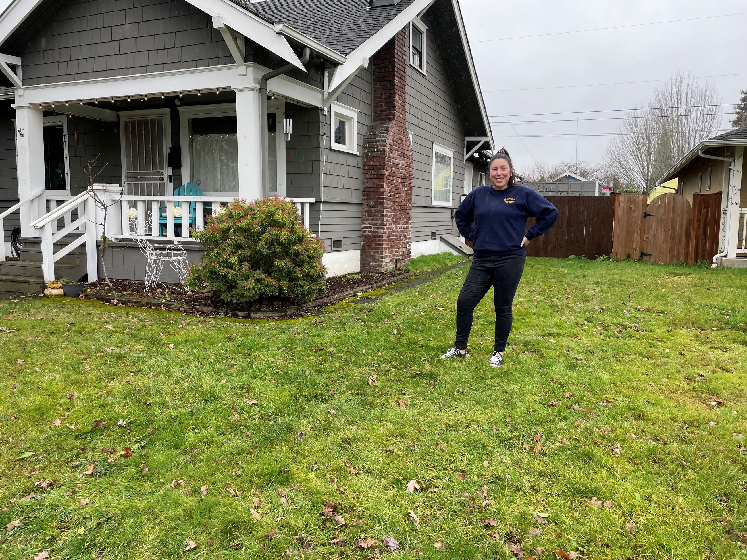 A woman stands in her lawn next to her 2 story house showing off what will become a raingarden.