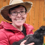 A person facing forward, holding a chicken with black feathers. They have a hat and j